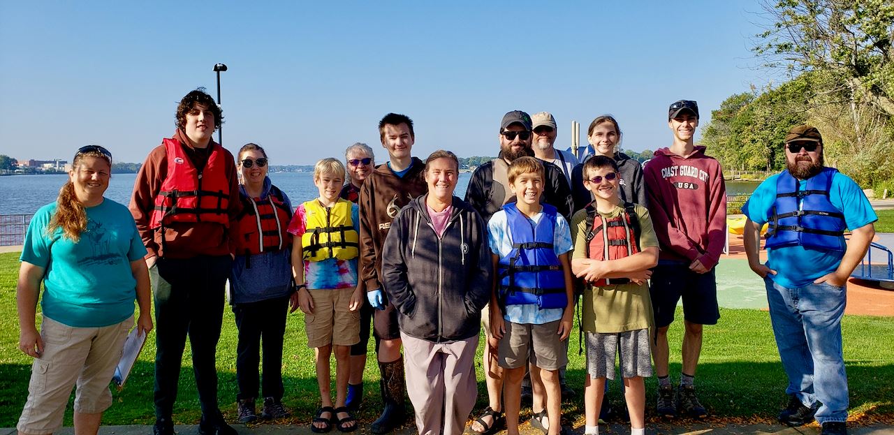 Group of 14 adults and youth standing in front of Lake, some wearing life jackets