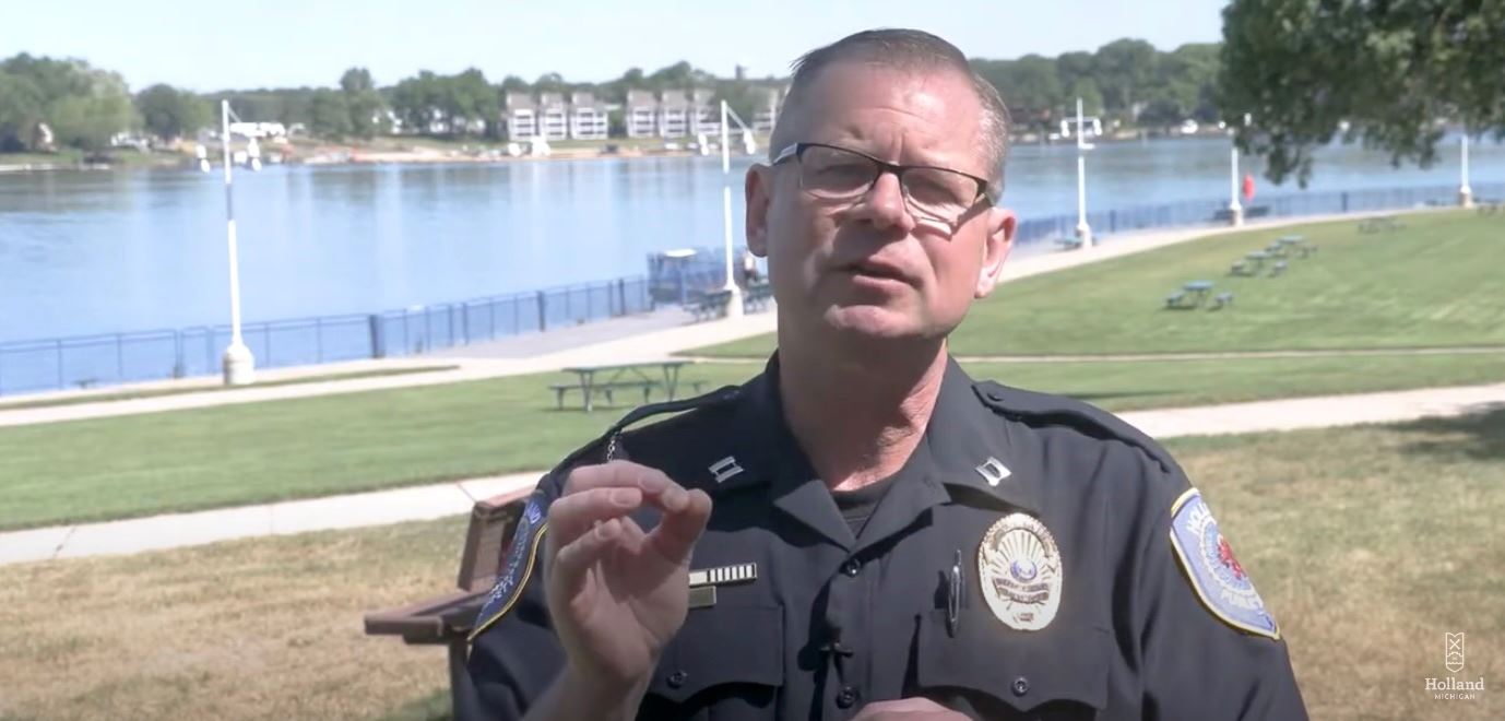 Police officer talking into camera, at park with a lake in the background, in the summer