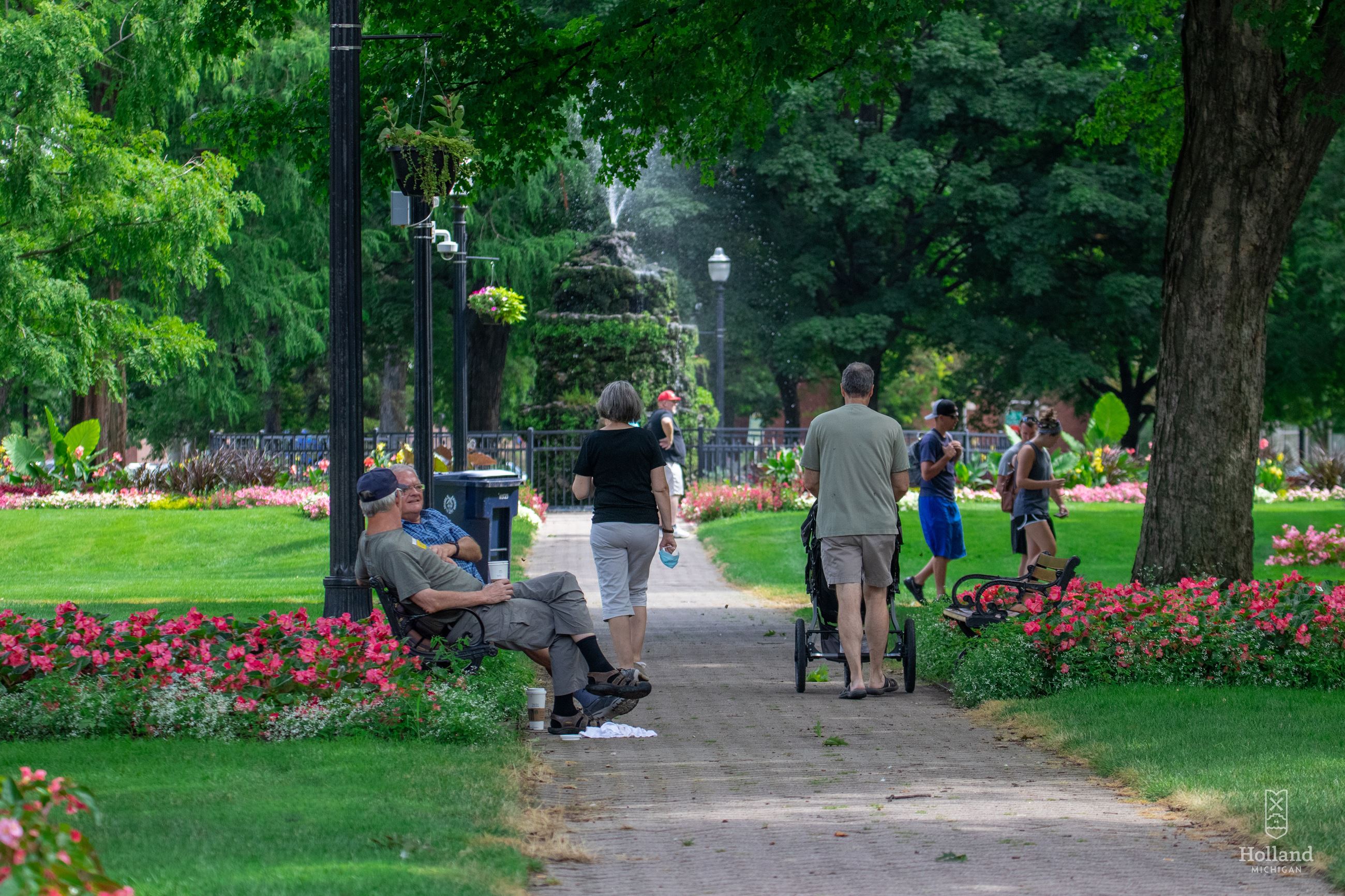 Summer day in a city park, 2 men sitting on a bench, a woman walking and man pushing a stroller. 