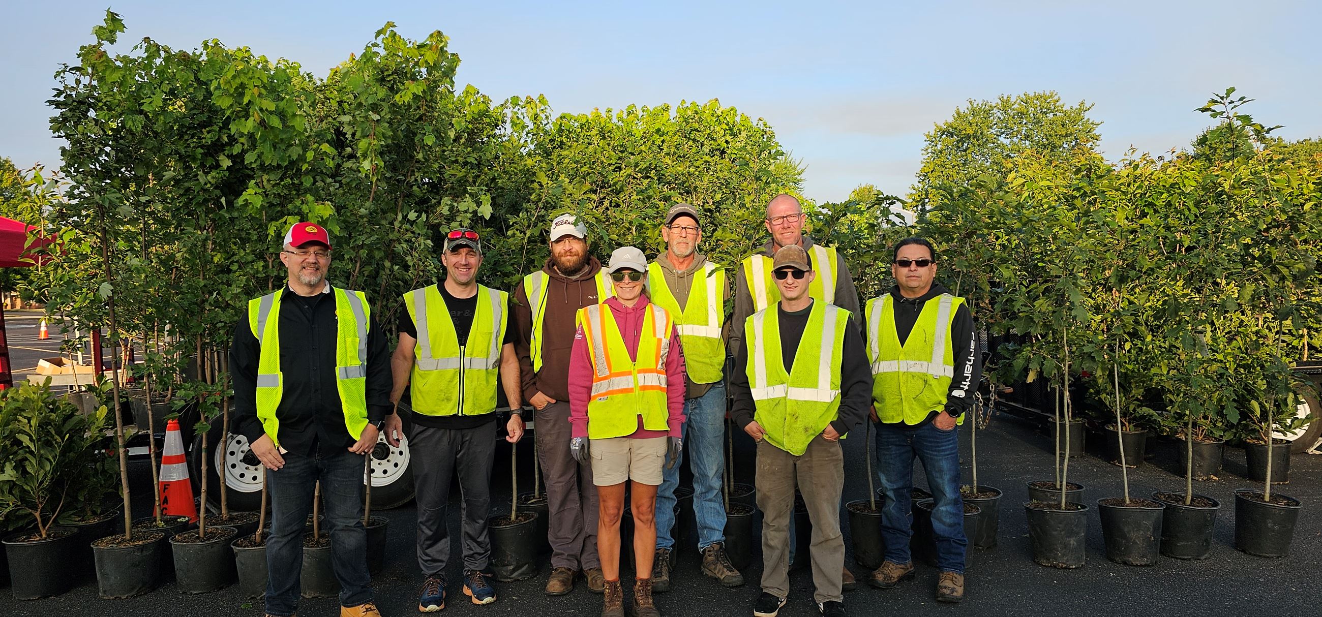 Eight men and women wearing fluorscent green vests stand in front of young trees in pots