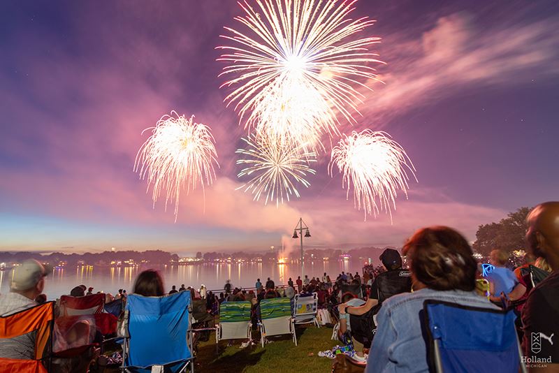 gathering of people seated on blankets at a park overlooking a lake watching fireworks