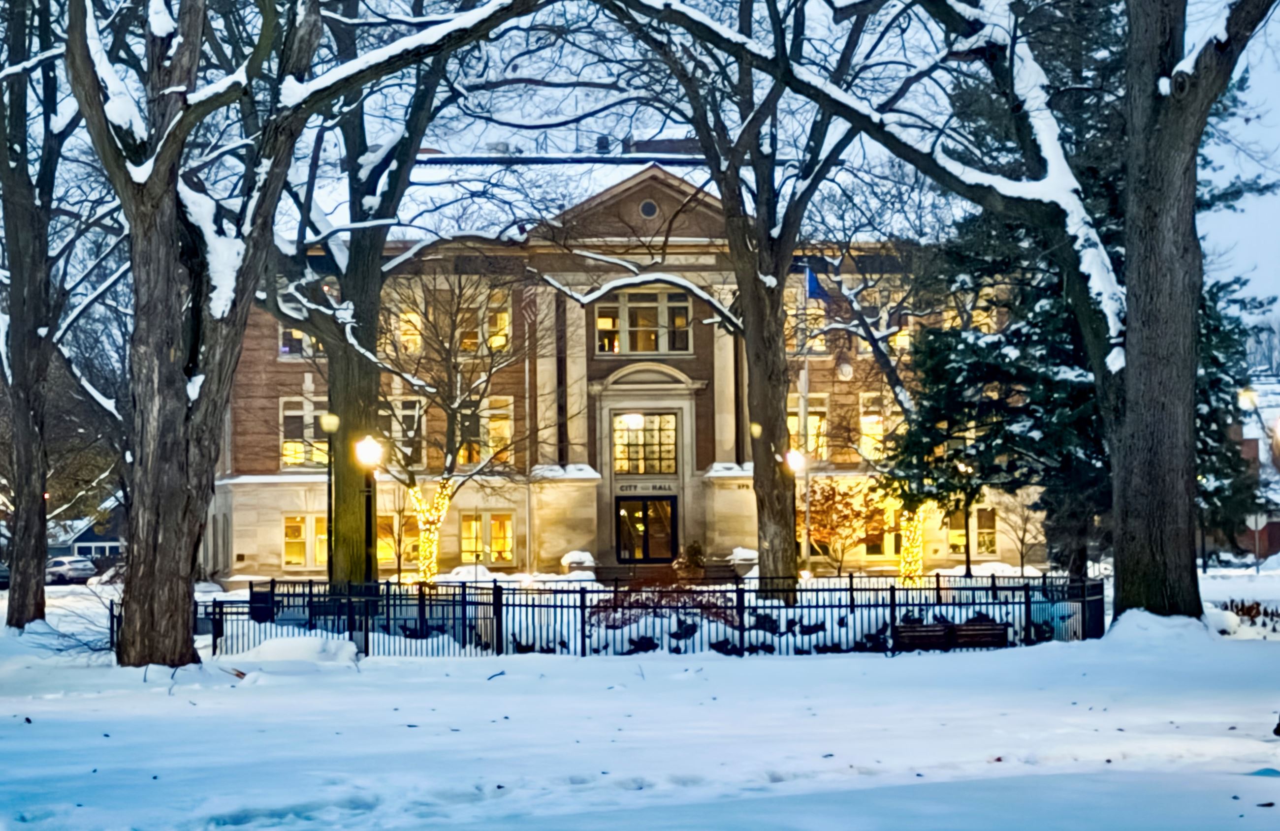 looking onto 3-story brick building through tress with Christmas lights and snow  
