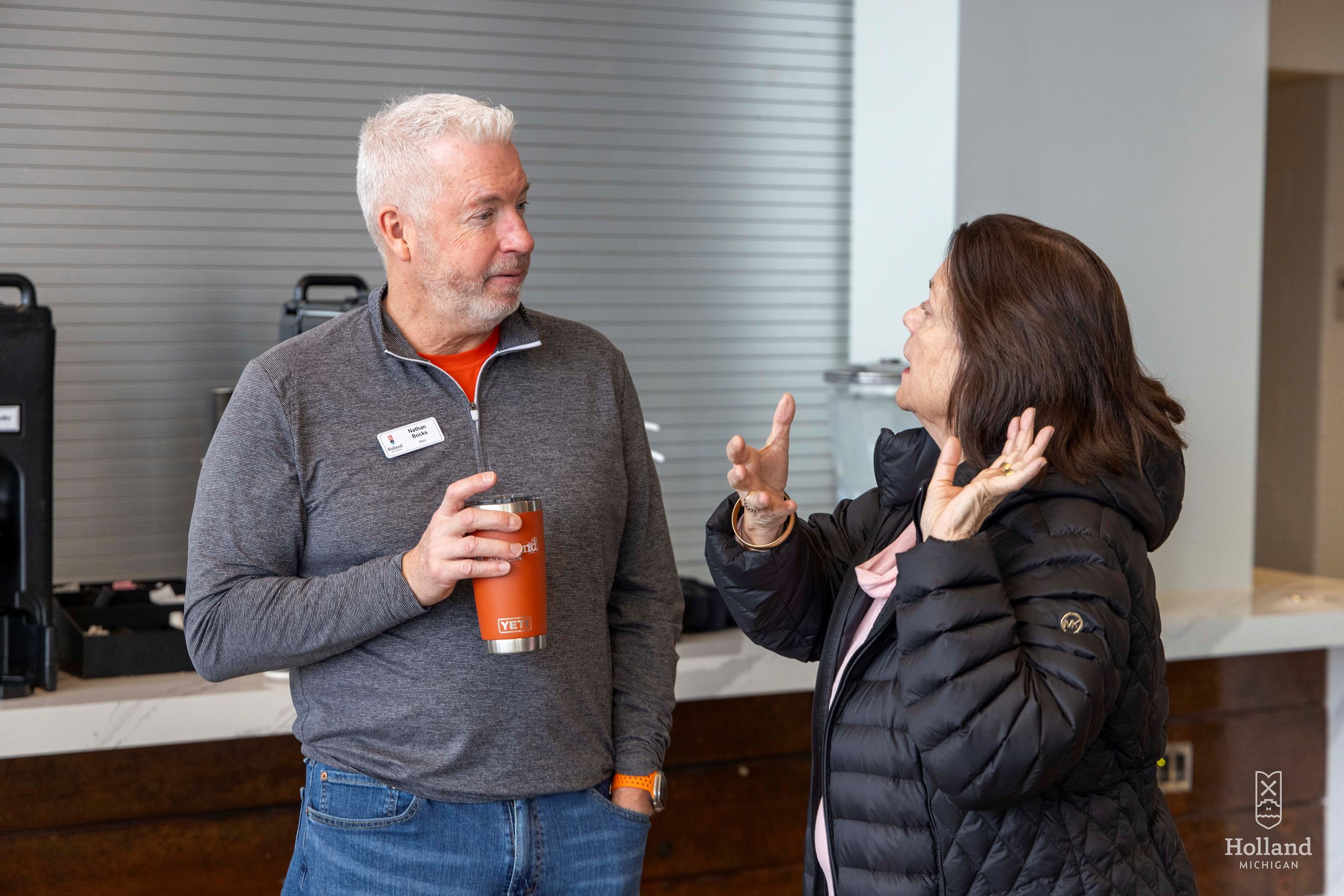 Man holding orange insulated cup talking with a woman inside a lobby of a civic center
