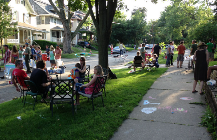 A group of neighbors sitting in chairs and mingling at a block party on a residential street.